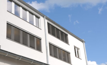 Ansicht eines Bürohauses mit Außenjalousien, teils ganz heruntergelassen – an einem Fenster hochgefahren