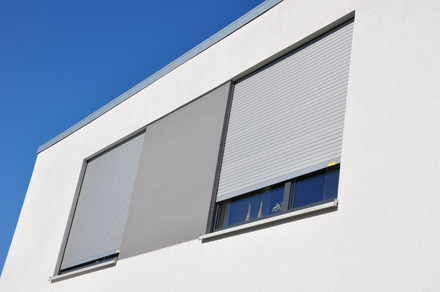 Außenansicht zweier Fenster eines Bürohauses mit heruntergelassenen Rollläden in lichtgrau
