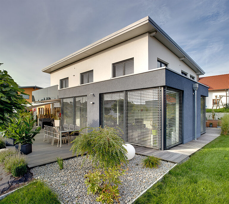 Zweistöckiges Wohnhaus mit einstöckigem Vorbau, dessen große Fensterfronten mit heruntergelassenen Außenjalousien beschattet sind. 