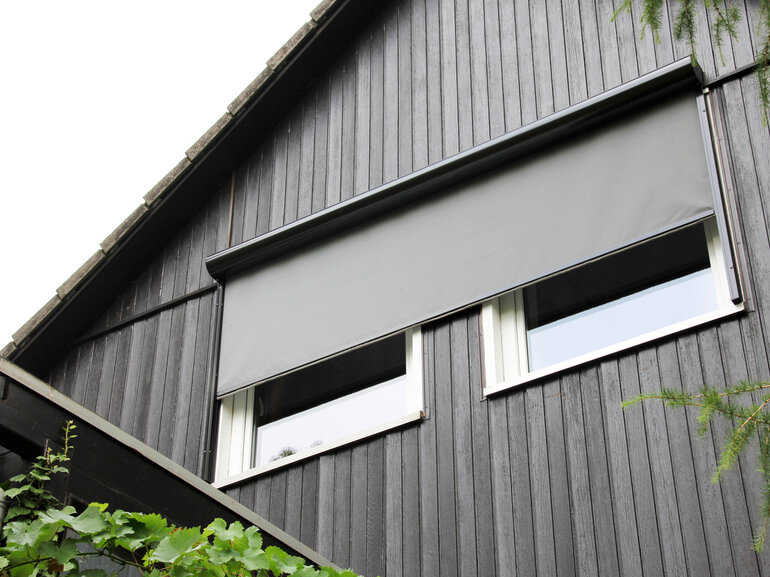 Heruntergelassene, nachträglich montierte Senkrechtmarkise in grau mit rundem Aluminiumkasten, die an einer Holzhausfront über zwei Fenster läuft